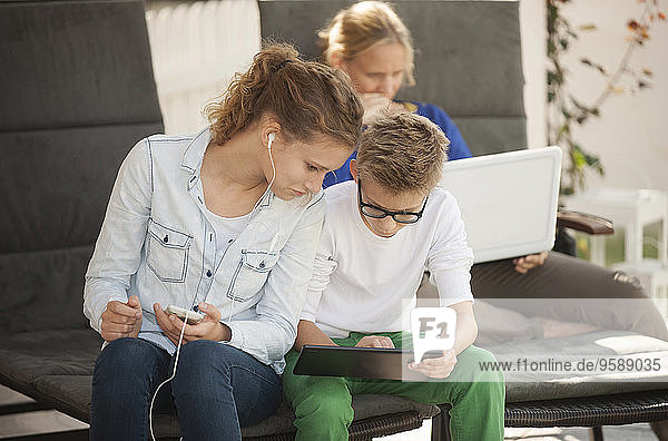 Mutter und ihre beiden Kinder entspannen sich mit digitalem Tablett  Smartphone und Laptop auf der Terrasse.