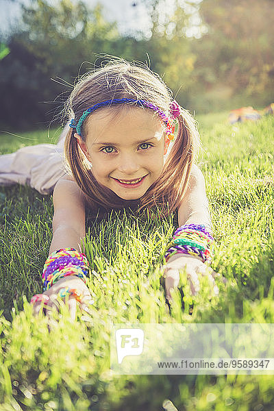 Porträt eines lächelnden kleinen Mädchens mit Webstuhlarmbändern und Haarband auf einer Wiese liegend
