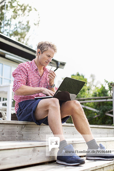Man using hands-free device and laptop while sitting on porch against clear sky