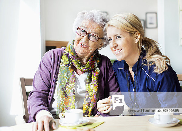Fröhliche Frau und Seniorin  die beim Sitzen am Tisch im Pflegeheim wegschaut.