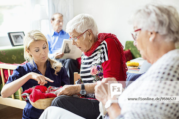 Female assisting senior women in knitting while man reading book in background at nursing home