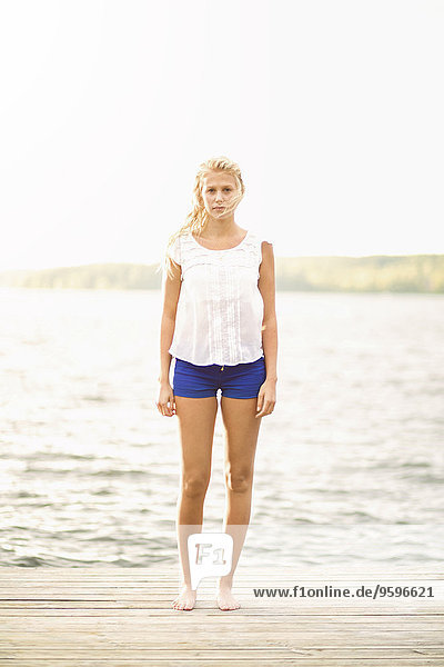 Ganzflächiges Porträt einer jungen Frau  die auf einer Strandpromenade am See steht.