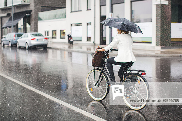 Durchgehende Rückansicht der Geschäftsfrau  die während der Regenzeit auf der nassen Stadtstraße Fahrrad fährt.