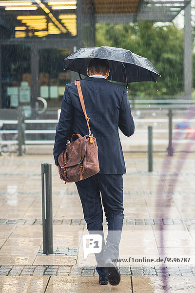 Rückansicht des Geschäftsmannes,  der während der Regenzeit auf dem Bürgersteig läuft.