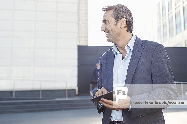 Smiling businessman looking away while using digital tablet outside office building