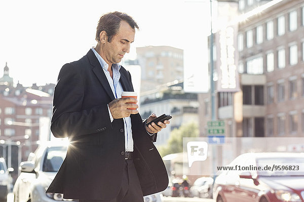 Businessman using mobile phone and holding disposable coffee cup while walking on city street