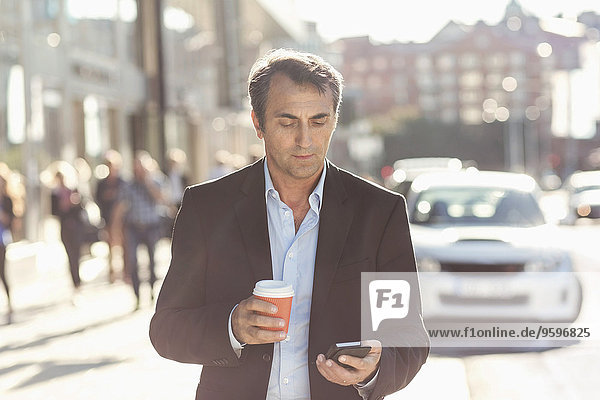 Geschäftsmann mit Handy und Einweg-Kaffeetasse beim Spaziergang auf der Stadtstraße