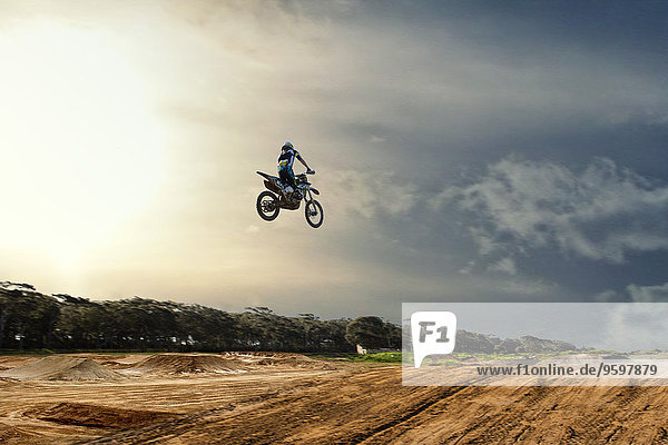 Silhouettierter junger  männlicher Motocross-Rennfahrer  der in der Luft über die Schlammbahn springt.