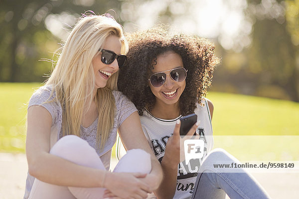 Zwei junge Frauen  die auf eine SMS auf dem Smartphone im Park herabblicken.