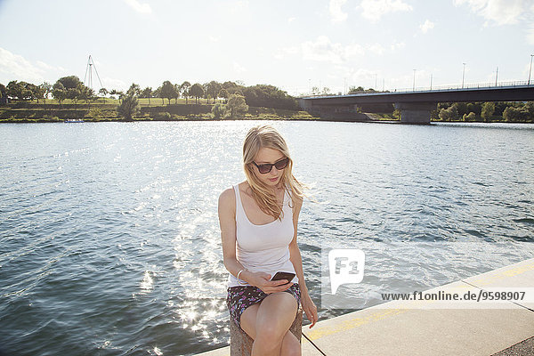Young woman sitting at riverside reading texts on smartphone  Danube Island  Vienna  Austria