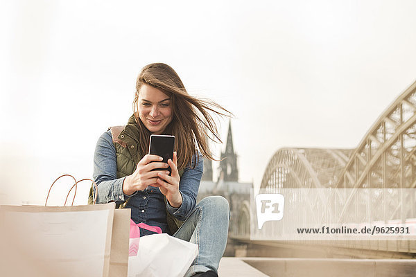 Deutschland  Köln  lächelnde junge Frau mit Einkaufstaschen  die einen Selfie nimmt