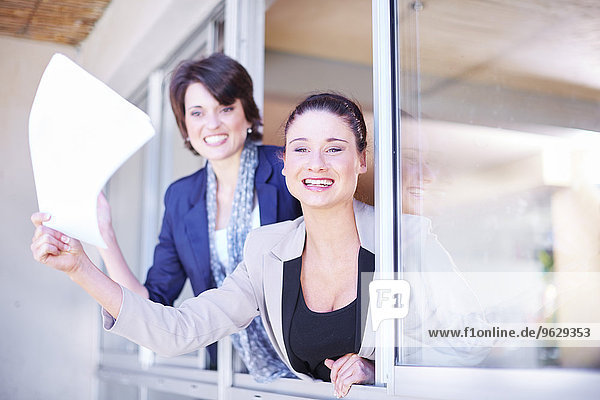 Two female creative professionals leaning out of window waving with sheet of paper