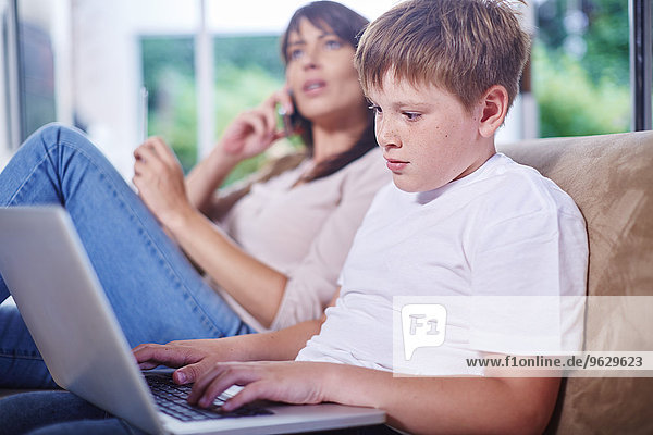 Junge zu Hause mit Laptop und Mutter im Hintergrund