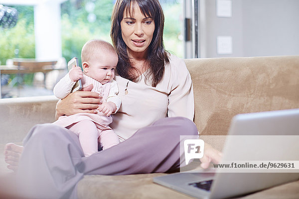 Mother sitting with baby on couch working on laptop