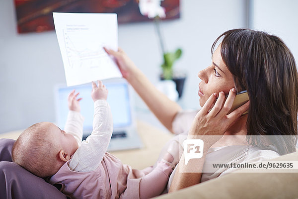 Mother with baby on couch talking on cell phone and looking at papers
