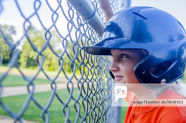 Junges Mädchen im Baseball-Kit  durch den Zaun schauend