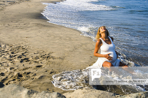 Schwangere reife Frau sitzt in Meereswellen am Strand  während sie den Bauch berührt.