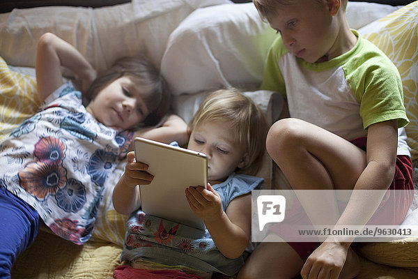 Drei kleine Kinder entspannen sich auf dem Bett mit Hilfe eines digitalen Tabletts.