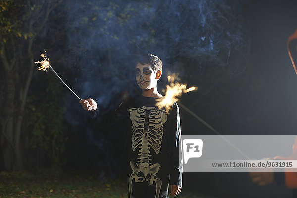 Junge im Halloween-Skelettkostüm mit Wunderkerze im Garten bei Nacht