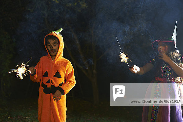 Geschwister in Halloween-Kostümen mit Wunderkerzen im Garten bei Nacht