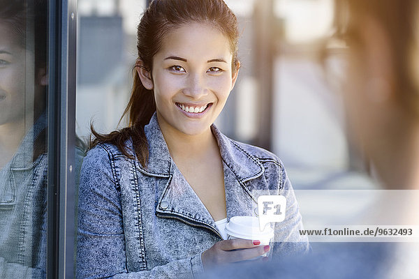Porträt einer jungen Frau  die Kaffee zum Mitnehmen trinkt und sich gegen das Parkgebäude lehnt.
