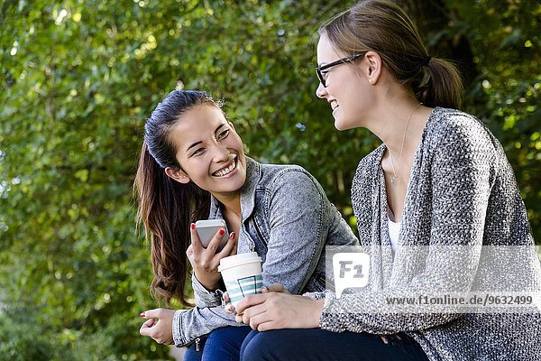 Two young women with takeaway coffee sitting chatting in park