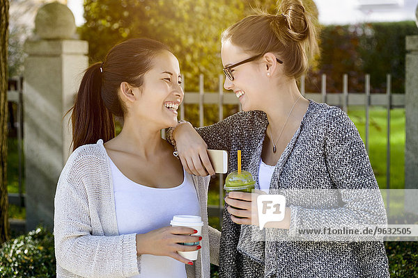 Zwei junge Freundinnen im Park  die lachen und Kaffee zum Mitnehmen trinken.