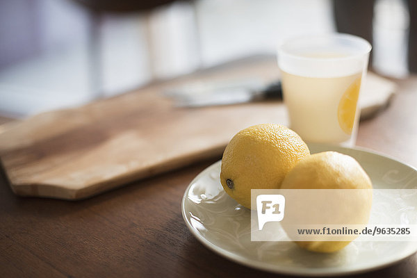 A chopping board with knife  a plate with two lemons and a glass on a table.