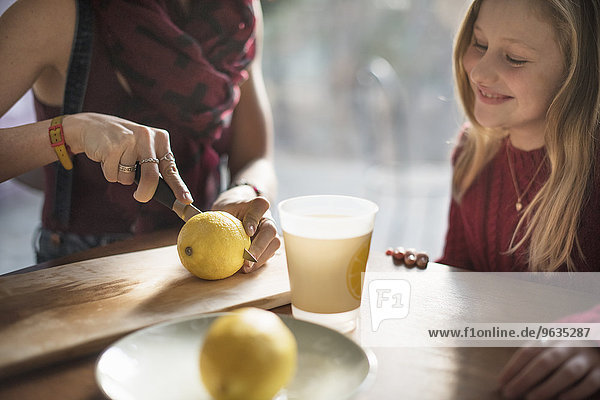 A woman and a smiling girl sitting at a table  woman slicing a lemon in half.