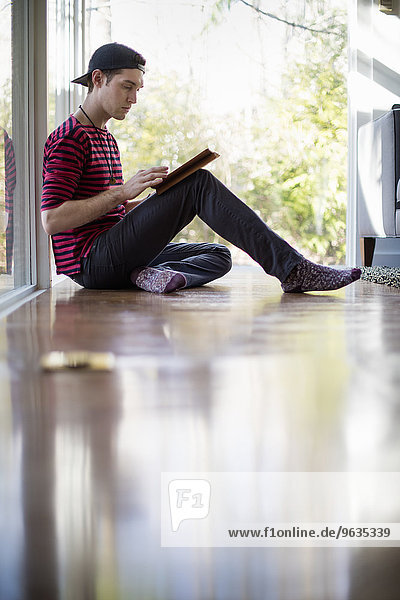 sitzend Mann sehen Boden Fußboden Fußböden Zimmer Mütze rückwärts Tablet PC Baseball Kleidung Wohnzimmer