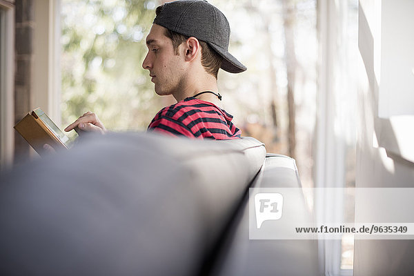 Man wearing a baseball cap backwards  sitting on a sofa  looking at a digital tablet.