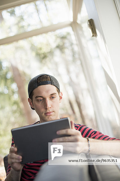 Man wearing a baseball cap backwards  sitting on a sofa  looking at a digital tablet.