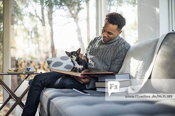 Man wearing a grey roll-neck jumper sitting on a sofa with a dog on his lap  looking at a book.
