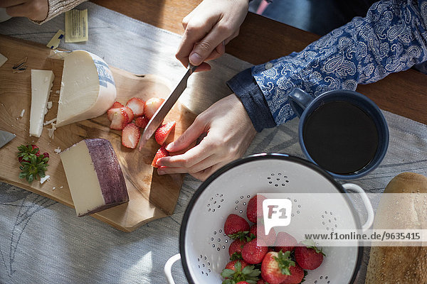 A woman slicing strawberries on a table with a wooden chopping board with a selection of cheeses and bread.