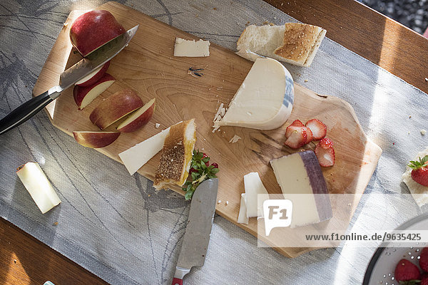 Knives and a wooden chopping board with a selection of cheeses  apples and bread.