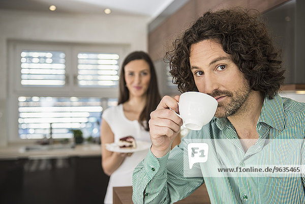 Man drinking cup of coffee and his wife with a cake in background