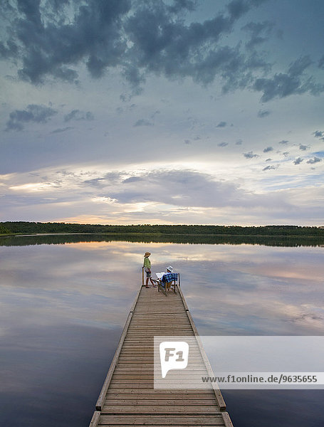 A couple  man and woman sitting at the end of a long wooden dock reaching out into a calm lake  at sunset.