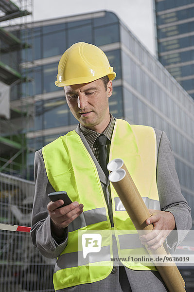 Site manager holding blueprints and text messaging on a mobile phone