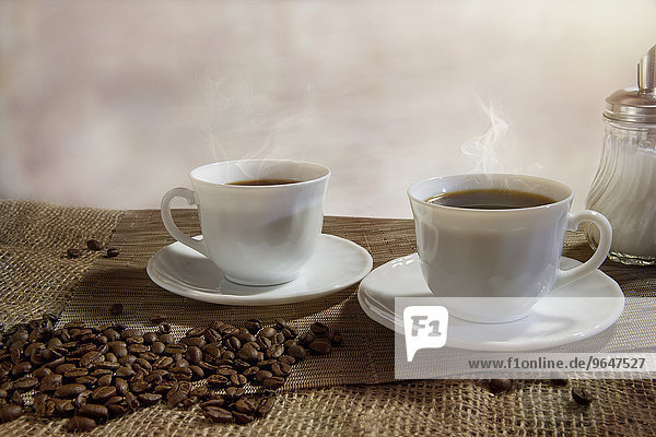 Zwei Tassen dampfender Kaffee  davor Kaffeebohnen
