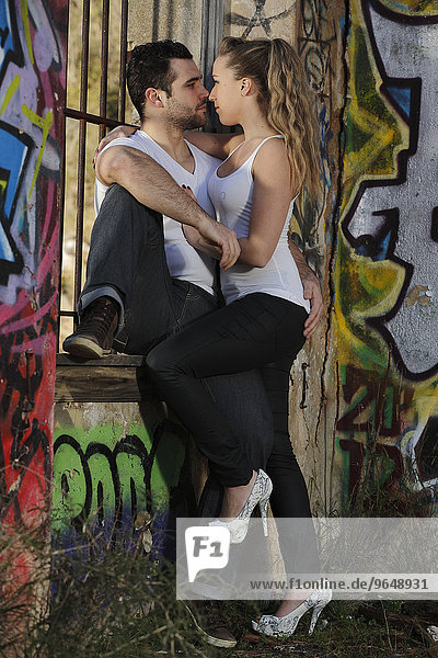Young couple kissing against a barred window in a ruined building covered in graffiti