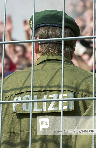 Polizist steht am Zaun im Fussballstadion und beobachtet die Fans