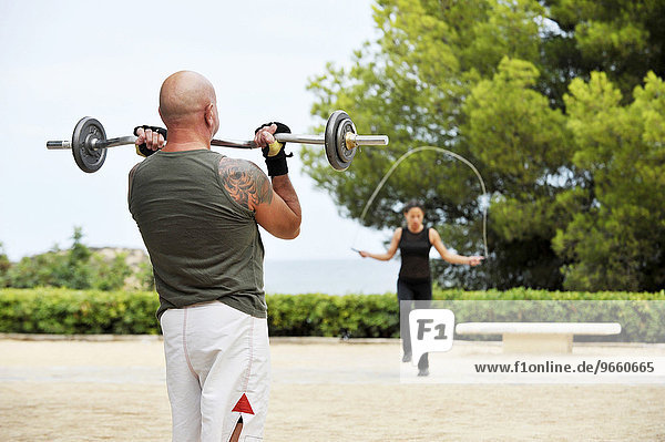 Ein Mann trainiert mit Gewichten während eine Frau Seil springt  in einem Park