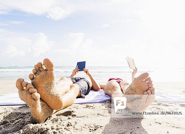 Close-up of sand-covered feet of young couple lying on blanket on beach reading