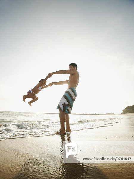 Mixed race father swinging daughter on beach