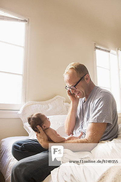 Father talking on cell phone and holding baby on bed