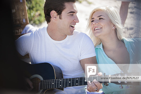 Junger Mann  der mit seiner Freundin am Strand Gitarre spielt.