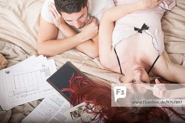Gelangweiltes Paar auf dem Bett liegend mit digitalem Tablett und Papierkram