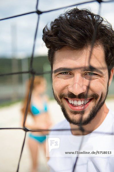 Porträt eines lächelnden jungen Mannes am Netz des Beachvolleyballfeldes