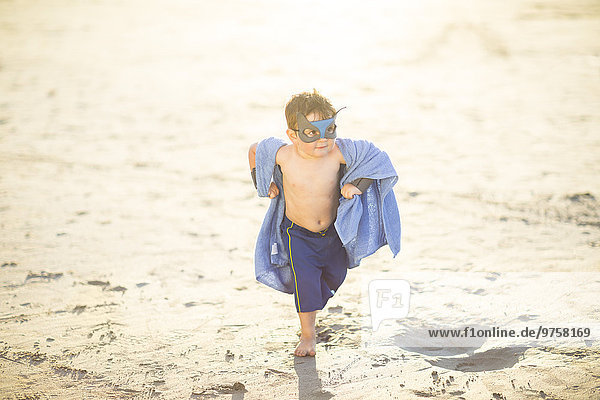 Kleiner Junge am Strand  verkleidet als Superheld mit Maske und Handtuch.