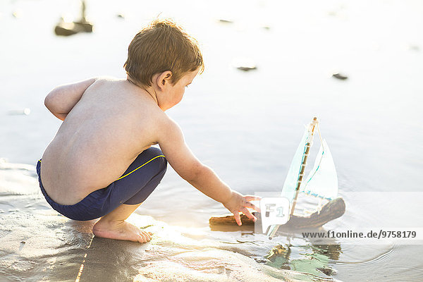 Junge am Strand spielt mit einem Spielzeug-Holzboot im Wasser